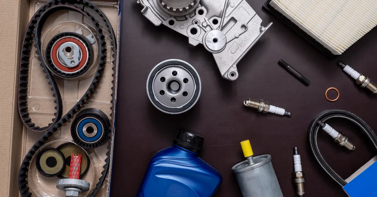A collection of car maintenance parts. Items include timing belts and pulleys in a cardboard box, a metallic engine component with gears, an oil filter, three spark plugs, a blue motor oil bottle, a cylindrical fuel filter with yellow caps, an air filter with pleated paper, and small bolts with a copper washer.