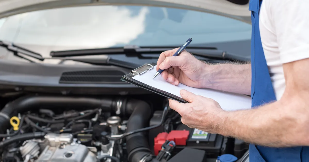 A person in blue overalls writing on a clipboard next to an open car hood.