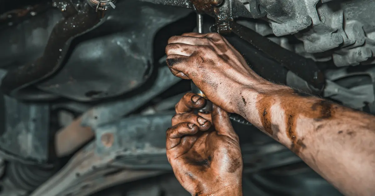 A close-up, darkly lit photograph of a person’s hands working on the underside of a vehicle engine.
