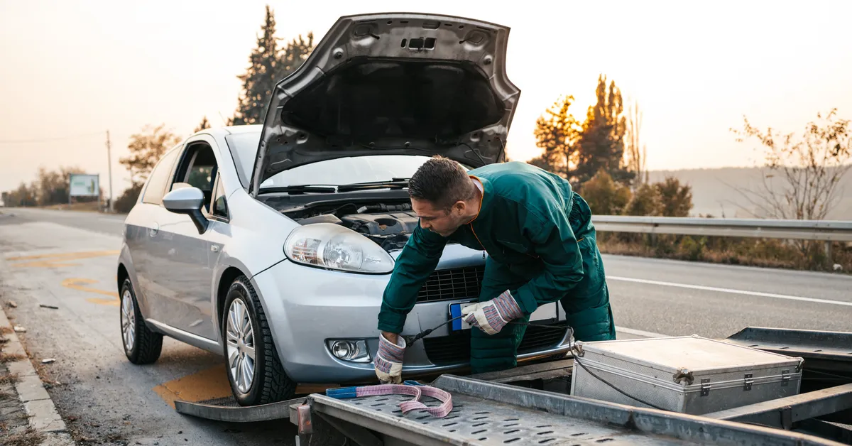 A man in a green jumpsuit working on a car with its hood open on the side of a road.