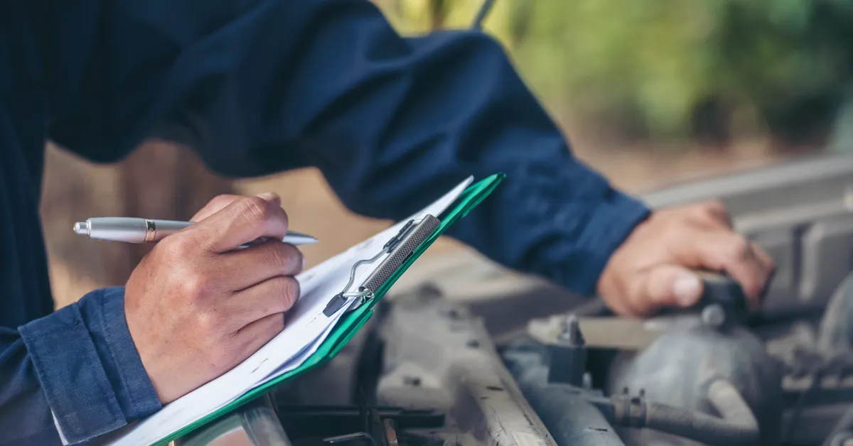 A person in a blue uniform holding a pen and a clipboard near a car engine.