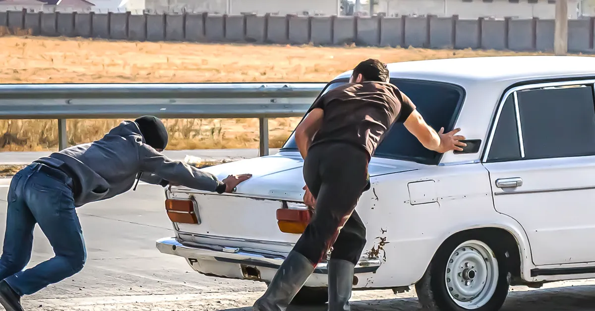 Two people in dark clothing pushing a white car on a road.