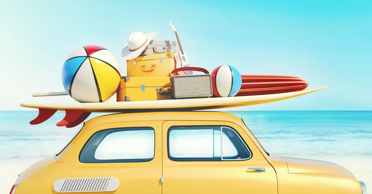 A yellow car parked by the beach. On the roof is a surfboard, beach ball, and suitcases.